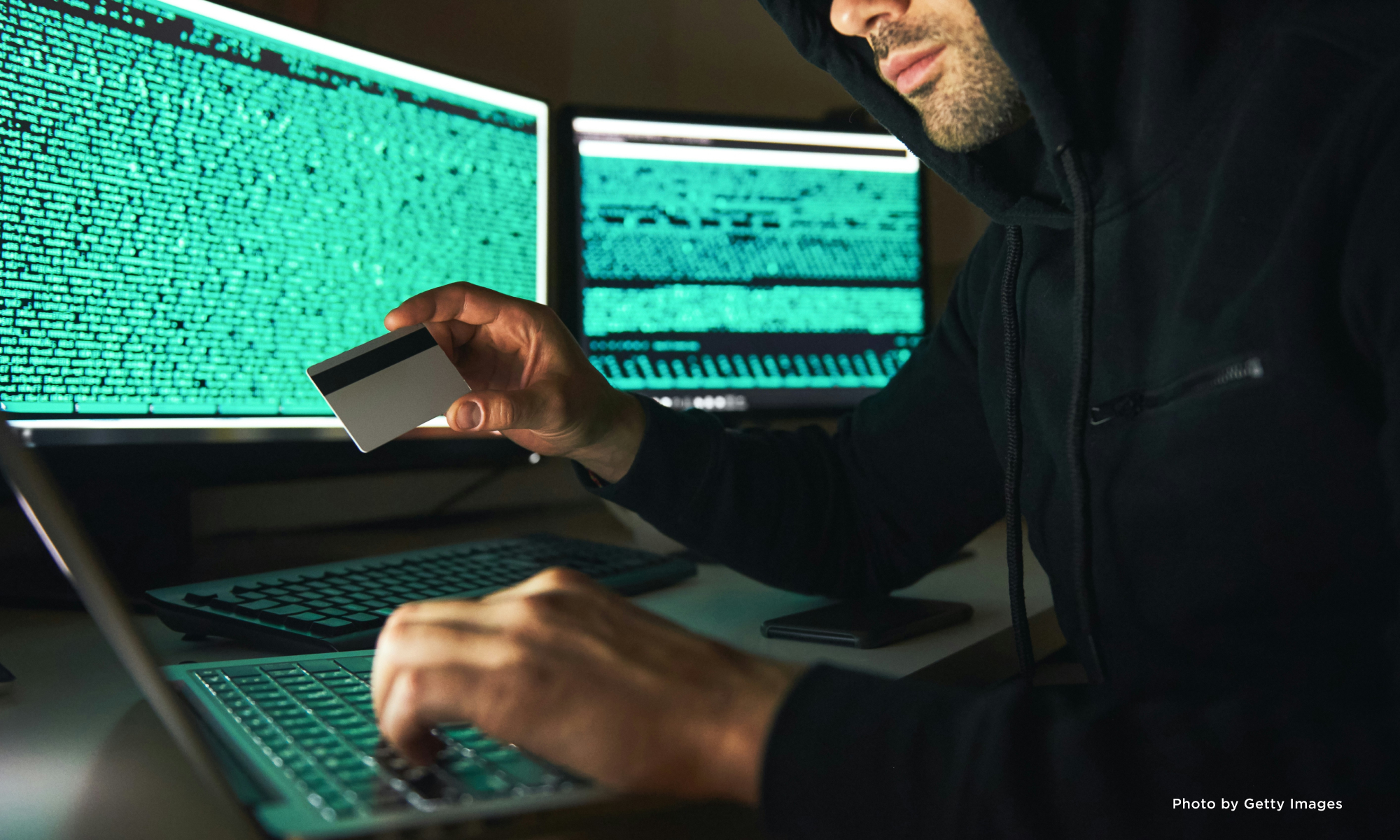 A hacker sitting in front of a computer screen holding a credit card.
