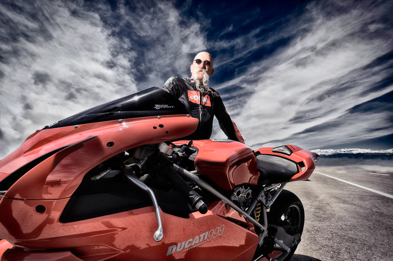 Dramatic photo of a man with a large moustache standing behind a sports motorcycle.