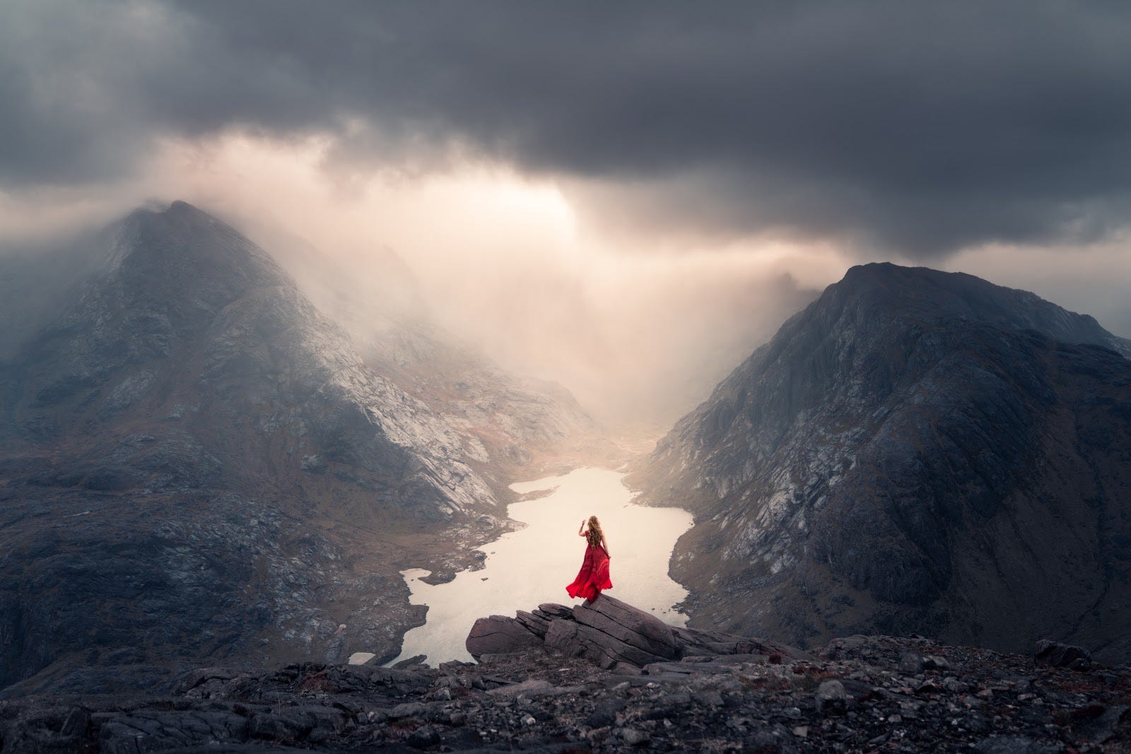 Woman in a red dress standing on a cliff overlooking a cloudy valley in Scotland.
