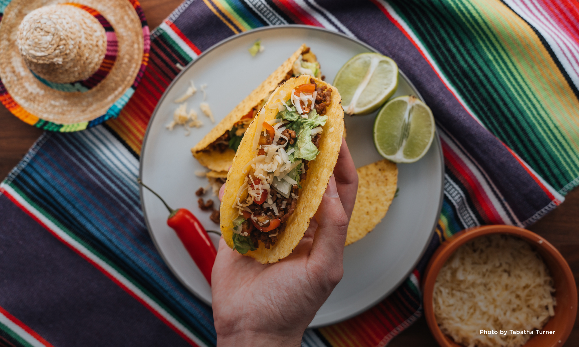 Overhead view of a hand holding a stuffed taco over a plate on a colorful tablecloth.