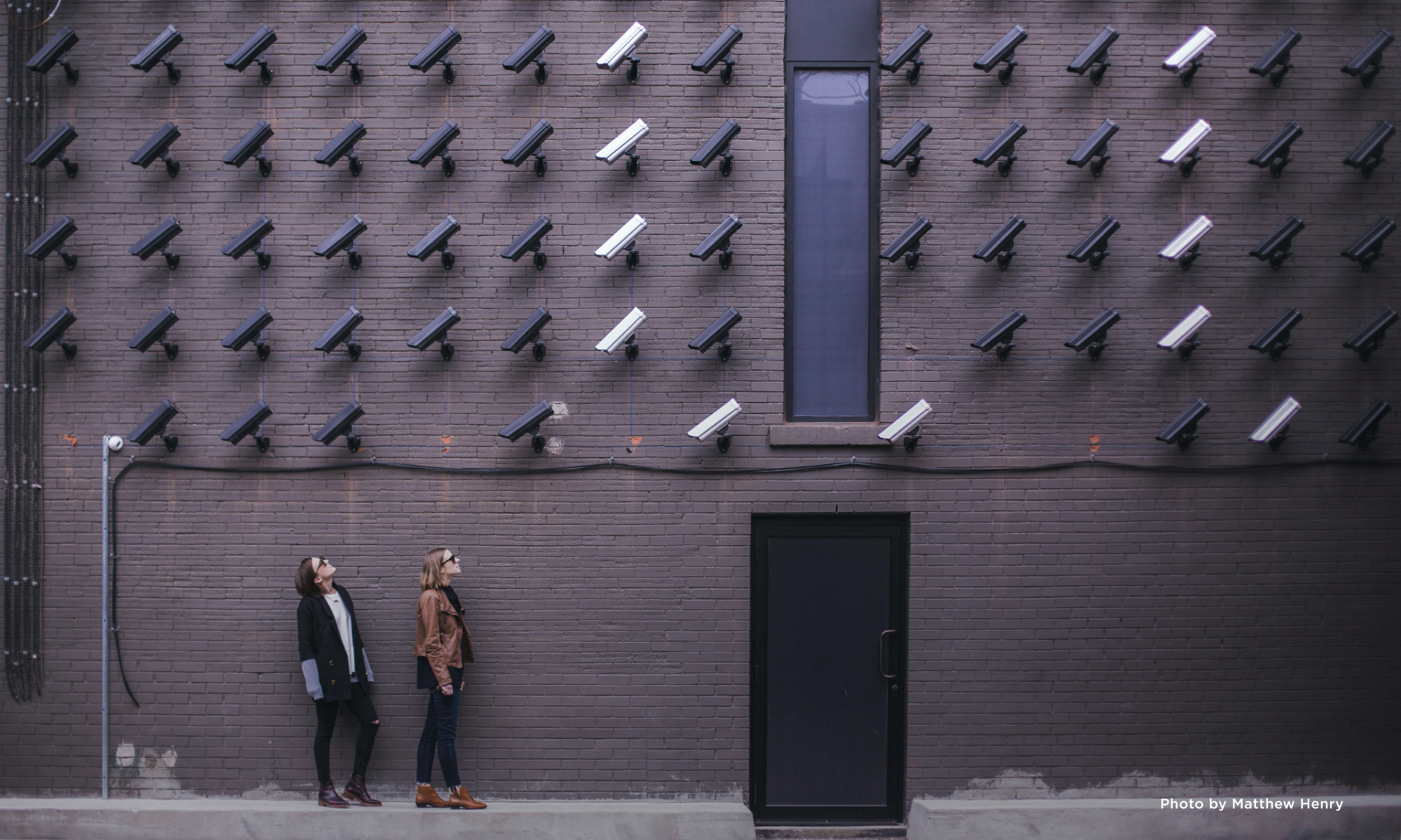 Two women standing in front of a brick wall filled with several security cameras pointed at them.