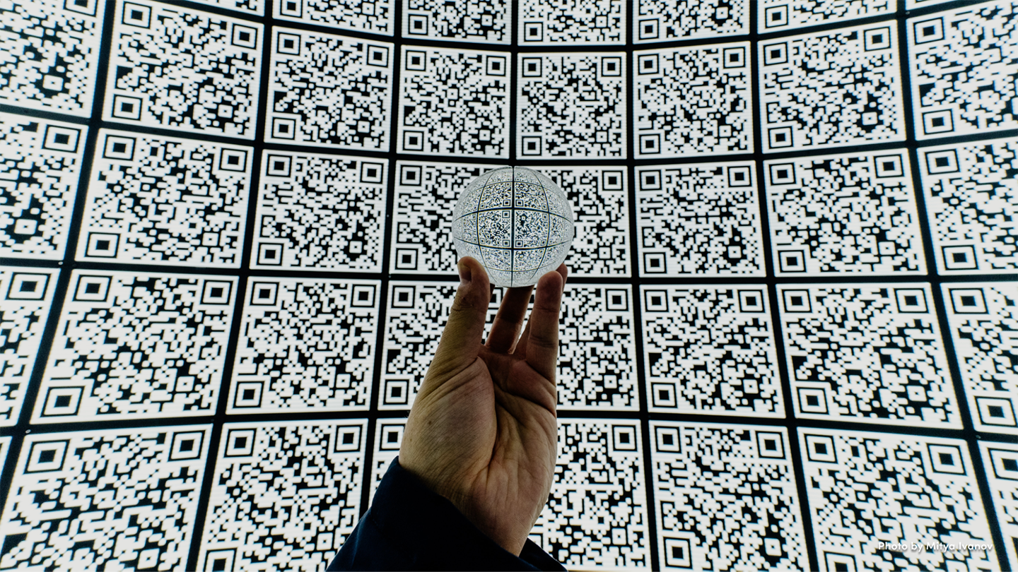 A hand holding a glass sphere in front of a curved screen filled with QR codes.